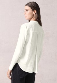 Langærmet hvid bluse med en afslappet pasform, silkeagtig tekstur og buet kant. Har en enkel krave og detalje med rygplissé.