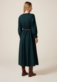 Dlouhé tmavě zelené šaty se staženým pasem, nafouklými rukávy a knoflíkovými manžetami. Kombinováno s hnědými kotníkovými botami. Hladká textura látky.