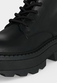 Botte en cuir noire avec semelle épaisse et rainurée et lacets ronds noirs passés dans des œillets en métal.