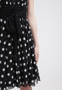 Robe noire à pois blancs, taille froncée et nœud noué ; bras droit visible à côté de la jupe sur un fond clair uni.