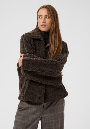 Vrouw met lang haar die een donkerbruine fleecetrui en geruite broek draagt, staat met gekruiste armen en kijkt opzij tegen een effen achtergrond.