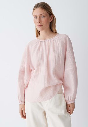Jonge vrouw die een losse, lichtroze blouse met textuur en lange mouwen draagt, gecombineerd met een witte broek, staand tegen een effen lichte achtergrond.