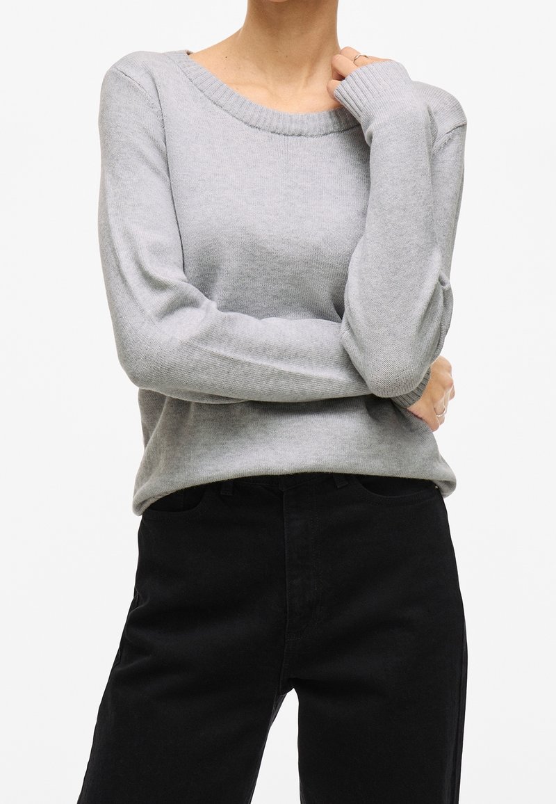 Personne portant un pull en maille à manches longues gris clair et un pantalon noir, bras croisés et une main touchant le cou.