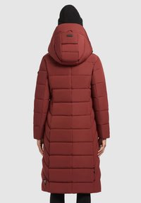Inimene kannab pikka burgundi värvi riiekattega talvejope, millel on kapuuts ja musta värvi beanie, vaadates seljaga lihtsalt taustast.