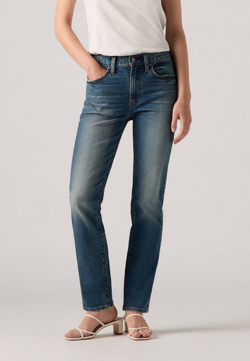 Femme portant un jean bleu taille mi-haute avec des zones délavées, un haut blanc sans manches et des sandales à talons beiges, debout avec une main dans la poche.