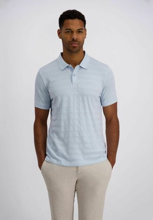 Mann mit kurzen lockigen Haaren, der ein hellblau gestreiftes Poloshirt und beige Hose trägt und mit den Händen in den Taschen vor einem schlichten Hintergrund steht.