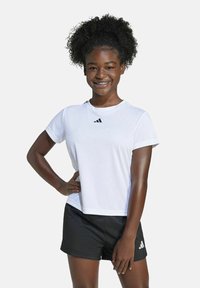Chica sonriente con cabello rizado que lleva una camiseta blanca de Adidas y pantalones cortos negros, de pie con una mano en la cadera frente a un fondo sencillo.