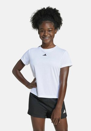 Chica sonriente con cabello rizado que lleva una camiseta blanca de Adidas y pantalones cortos negros, de pie con una mano en la cadera frente a un fondo sencillo.