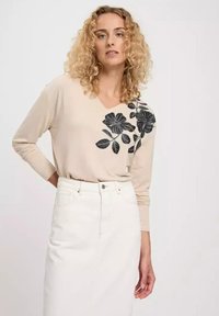 Beżowa bluzka z długim rękawem i dekoltem w serek, z czarnym kwiatowym nadrukiem po jednej stronie, zestawiona z białą spódnicą dżinsową. Gładka tekstura materiału.