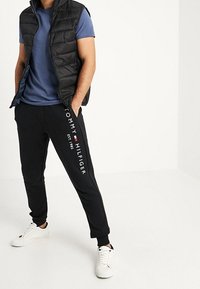 Czarna pikowana kamizelka, niebieski T-shirt, czarne spodnie dresowe z białym nadrukiem "TOMMY HILFIGER" oraz białe trampki. Gładki materiał, luźny krój.