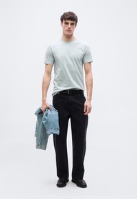 Νέος άνδρας όρθιος, φοράει ανοιχτό πράσινο μπλουζάκι, μαύρο παντελόνι και παπούτσια, κρατώντας ένα διπλωμένο ελαφρύ μπλε τζιν μπουφάν στο αριστερό του χέρι.