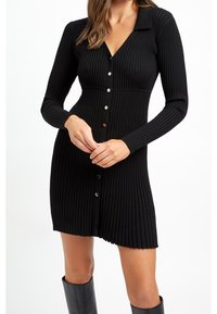 Abito nero in maglia a coste con maniche lunghe, scollo a V e chiusura frontale con bottoni. Presenta un corpetto aderente e un orlo svasato.