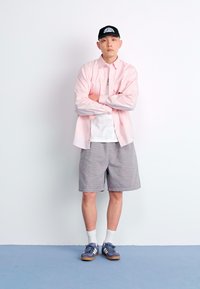 Camisa de botão rosa clara sobre uma t-shirt branca, calções cinzentos, meias brancas e ténis azuis. Boné preto com logótipo branco. Braços cruzados.