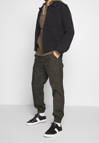 Černá džínová bunda, olivově zelená mikina s kapucí, tmavě zelené nákladní kalhoty a černé tenisky s bílými doplňky. Příležitostné kapsy na kalhotách.