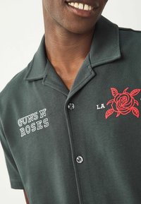 Krátký rukáv, tmavě zelená košile z měkkého materiálu, se bílým textem "GUNS N' ROSES" a designem červené růže na levé straně hrudi. Černé zapínání na knoflíky.