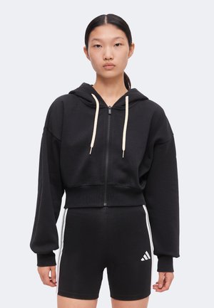 Mladá žena má na sebe čiernu krátku mikinu na zips s bielymi šnúrkami a čierne šortky s bielymi pásikmi a malým logom na nohe.