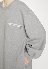 Ljusgrå överdimensionerad sweatshirt gjord av mjukt tyg, med en bröstficka med fransar och ribbade muddar.