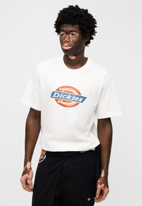 Άσπρο βαμβακερό T-shirt με μπλε και πορτοκαλί λογότυπο "Dickies" στο μπροστινό μέρος. Το μοντέλο φοράει μαύρο παντελόνι, εμφανίζοντας ελαφρές δερματικές ατέλειες στα χέρια.