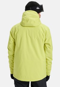 Heldere gele waterdichte jas met capuchon en lange mouwen, met een naadloos ontwerp en een gladde textuur. Zwarte handschoenen zichtbaar.