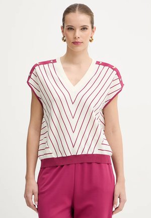 Donna con top bianco senza maniche a righe verticali rosa che formano un motivo a chevron e pantaloni rosa abbinati, in piedi davanti a uno sfondo bianco.