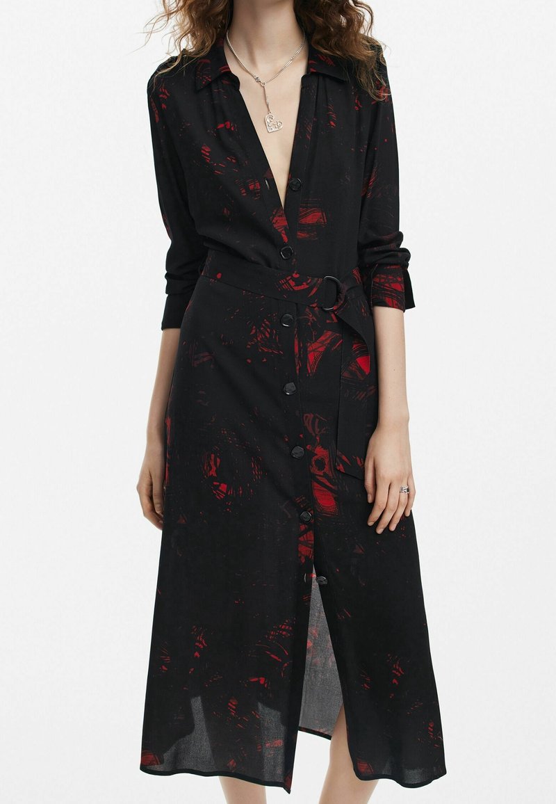 Femme portant une robe midi noire à motifs abstraits rouges, avec boutonnage sur le devant, ceinture à boucle à la taille, et manches trois-quarts, debout.