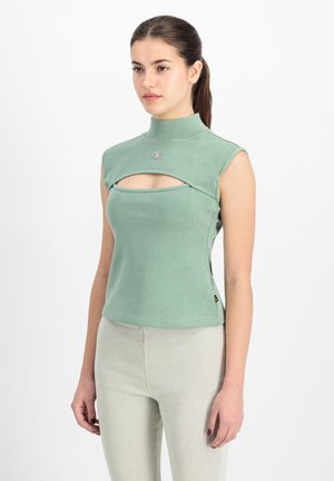 Kobieta o ciemnych włosach związanych w kucyk, ubrana w bezrękawnik w zielonym kolorze z wycięciem na klatce piersiowej i jasne, dopasowane spodnie, stojąca na białym tle.