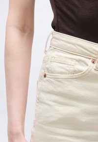 Άτομο που φοράει σκούρο καφέ τοπ και παντελόνι από κρεμ denim με ψηλή μέση και χάλκινα καρφιά, εμφανίζοντας τον κορμό και το αριστερό χέρι σε φόντο μονόχρωμο.