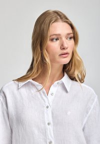 Chemise en lin blanc avec un devant à boutons, arborant un petit emblème sur la poitrine gauche et un col classique. Texture douce avec une coupe décontractée.