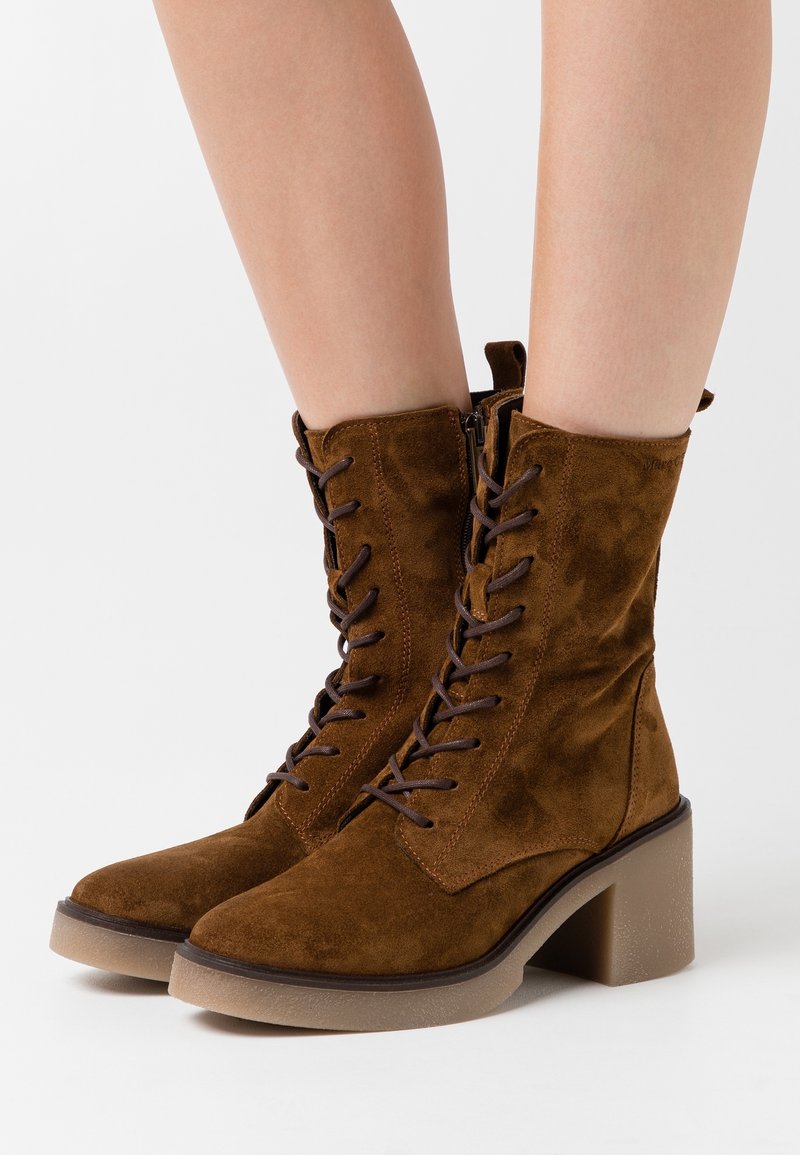Bottines à lacets en daim marron avec talons épais portées sur des jambes nues sur un fond blanc.