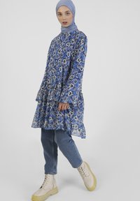 Kwiatowa sukienka w odcieniach niebieskiego, wykonana z lekkiego materiału, z falbaniastym dołem, noszona na niebieskich dżinsach i beżowych butach na sznurówki. Długie rękawy, wysoki dekolt.