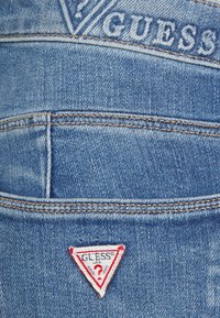 Tela de denim azul con una textura desgastada. Presenta un logo blanco y una etiqueta triangular con borde rojo que contiene detalles de marca y un signo de interrogación.