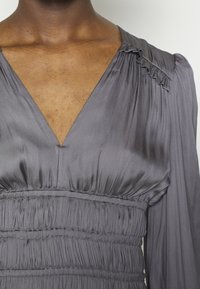 Robe en satin gris avec un décolleté en V profond, des détails froncés à la taille et des manches longues avec de subtils accents de volants. Texture lisse.