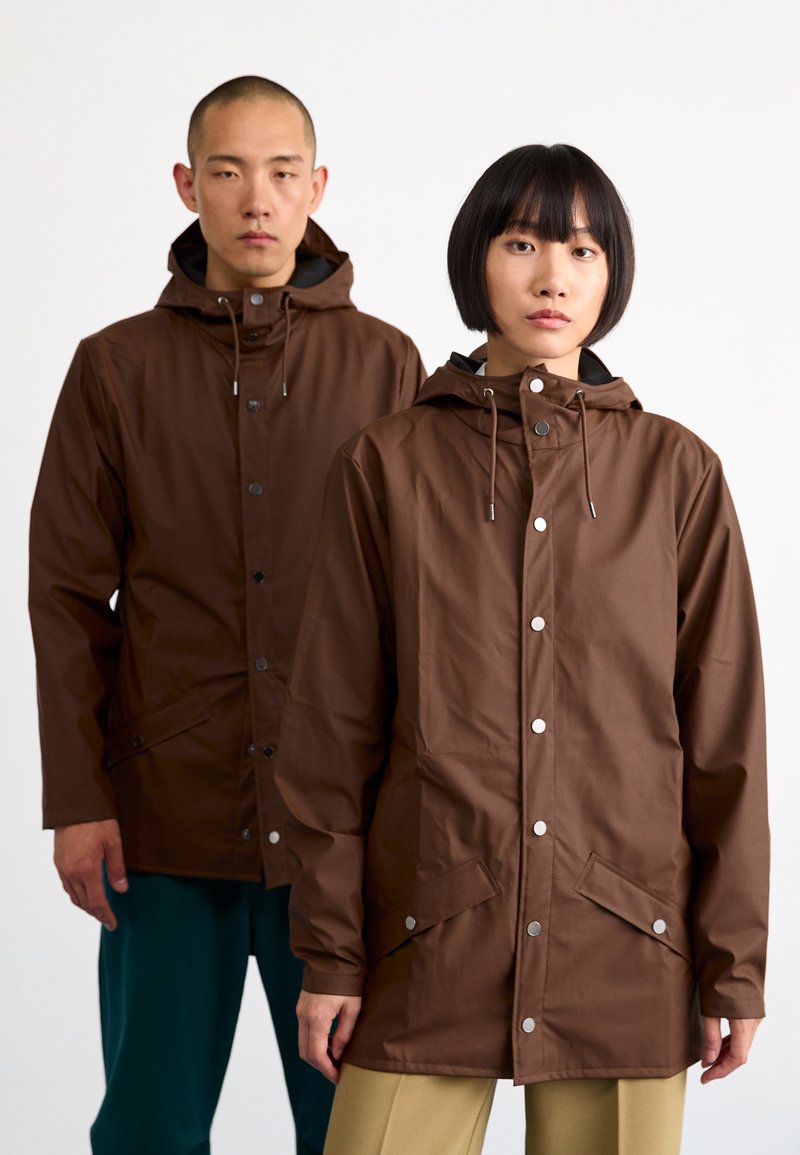 Vêtements imperméables marron avec capuches, dotés de fermetures à pression et de poches avant, portés par deux personnes sur un fond uni.