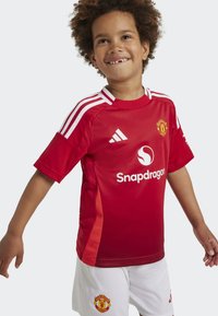 Maillot rouge Adidas de Manchester United avec des rayures blanches, logo sur la poitrine et accent rouge sur les manches. Porté avec un short blanc arborant l'écusson de l'équipe.