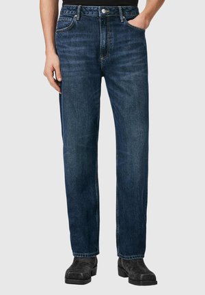 Person trägt dunkelblaue Jeans mit geradem Bein, Vorderentaschen, Knopf- und Reißverschluss sowie schwarzen Wildlederstiefeln und steht vor einem einfarbigen Hintergrund.