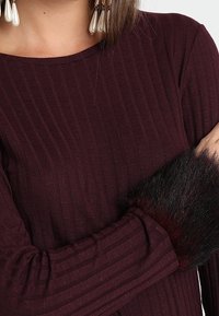 Vrouw in een ribbed, lange mouw, donker bordeaux shirt met zwarte nep bont manchetten en lange parel druppel oorbellen.