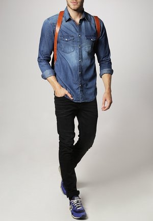 Camicia di jeans con due tasche sul petto, maniche arrotolate e spalline marroni in pelle. Pantaloni neri aderenti e sneakers blu completano l'outfit.