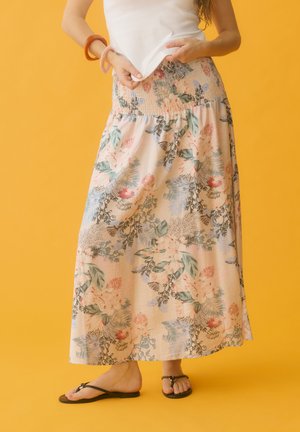 Femme portant une longue jupe à motif floral, un haut blanc sans manches, des sandales noires et deux bracelets, debout devant un fond jaune.