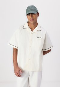 Short-sleeve, light beige shirt with black piping and a chest pocket featuring "Stan Ray" embroidery. Paired with a green cap.