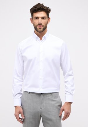 Chemise blanche en coton avec un col à boutons, des manches longues et une texture lisse, portée avec un pantalon gris clair.