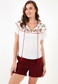 Witte blouse met geborduurd bloemenontwerp in rood en groen, korte mouwen en een V-hals. Heeft een verstelbare koord met rode kwastjes.