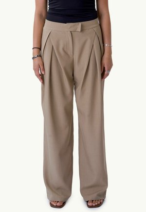 Femme portant un pantalon beige à jambes larges, un haut noir, des sandales et des bracelets en argent, debout les bras détendus le long du corps sur un fond uni.