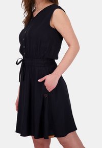 Abito nero senza maniche con bottoni sul davanti, vita svasata, tasche e una piccola patch a forma di cuore. Realizzato in tessuto morbido e leggero.