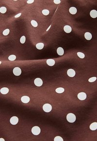 Ruskea kangas valkoisilla pilkuilla, pehmeä tekstuuri ja kevyet laskokset, jotka luovat varjoja ja korostuksia pinnalle.