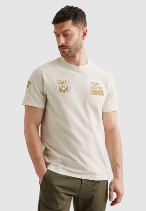 Mann trägt ein beigefarbenes PME Legend Kurzarmhemd mit gestickten Logos, grüne Hose, steht mit einer Hand in der Tasche.