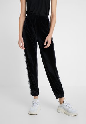 Persoon draagt zwarte velours joggers met witte zijstrepen en witte chunky sneakers, staand tegen een effen lichte achtergrond.