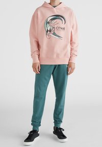 Roze hoodie met een grafische print van een surfs cène, gecombineerd met groene joggingbroek en zwarte sneakers met witte zolen.