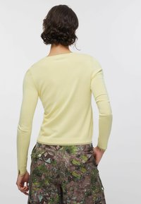 Rövid, göndör hajú személy halvány sárga hosszú ujjú felsőt és terepmintás zsebes nadrágot visel, egyik kezét a zsebében tartva áll.