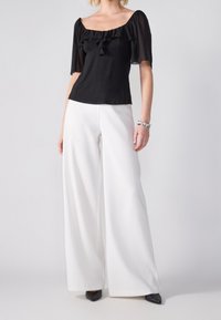 Černá blůza s výstřihem do ramen a volánkovými detaily, zkombinovaná s bílými širokými kalhotami a černými lodičkami na špičce.
