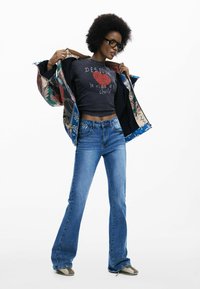 Jeans di jeans svasati, t-shirt grafica scura, giacca con motivi multicolori e accenti floreali. Occhiali con montatura nera e capelli ricci e corti.